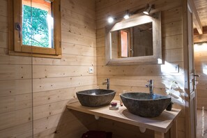 A wooden cabin bathroom with two large stone sinks, a mirror, and a window with a view of trees.