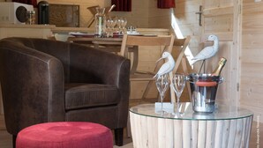 A cozy room with a leather armchair, a round ottoman, a glass table with a metal bucket containing a bottle and glasses, and a wooden wall. - LUXURY CABIN WITH SPA IN TREES (La Bussiere)