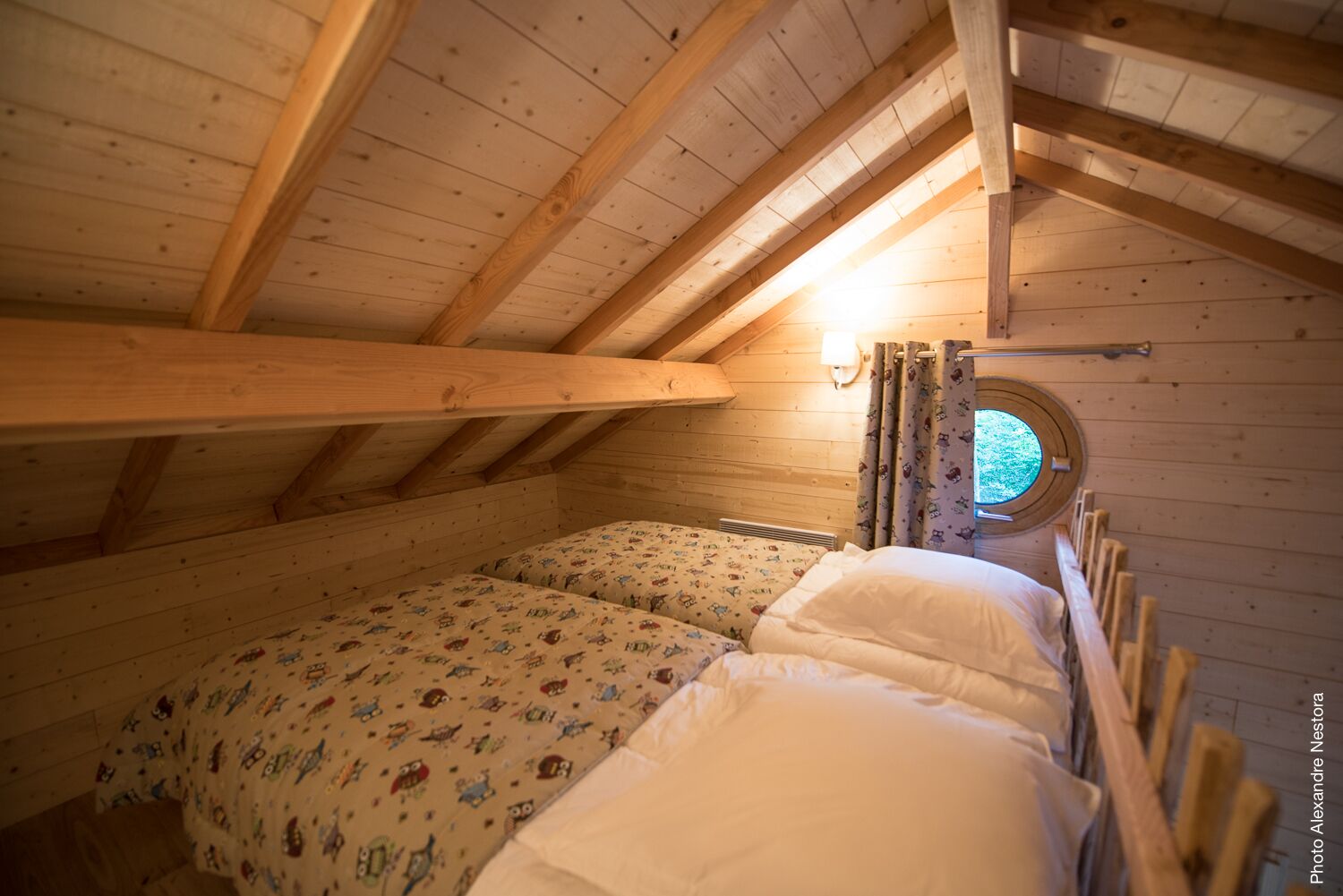 Ein gemütliches Schlafzimmer im Dachgeschoss mit Holzbalken, einem Rundfenster und zwei Betten mit gemusterten und schlichten Betttextilien.