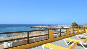 Ein Balkon mit gelben und weißen Liegestühlen mit Blick auf eine felsige Küstenlinie, Meer und eine ferne Stadtansicht.