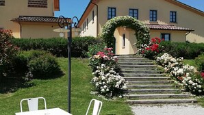 A patio area with white metal chairs and a table, a garden with steps leading to a house, and a clear blue sky. - SANGIOVESE APARTMENT at Agriturismo MUSIGNANO (Cerreto guidi)