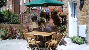 Um pátio com mesa e cadeiras de madeira, um guarda-sol verde e cestos de flores pendurados.