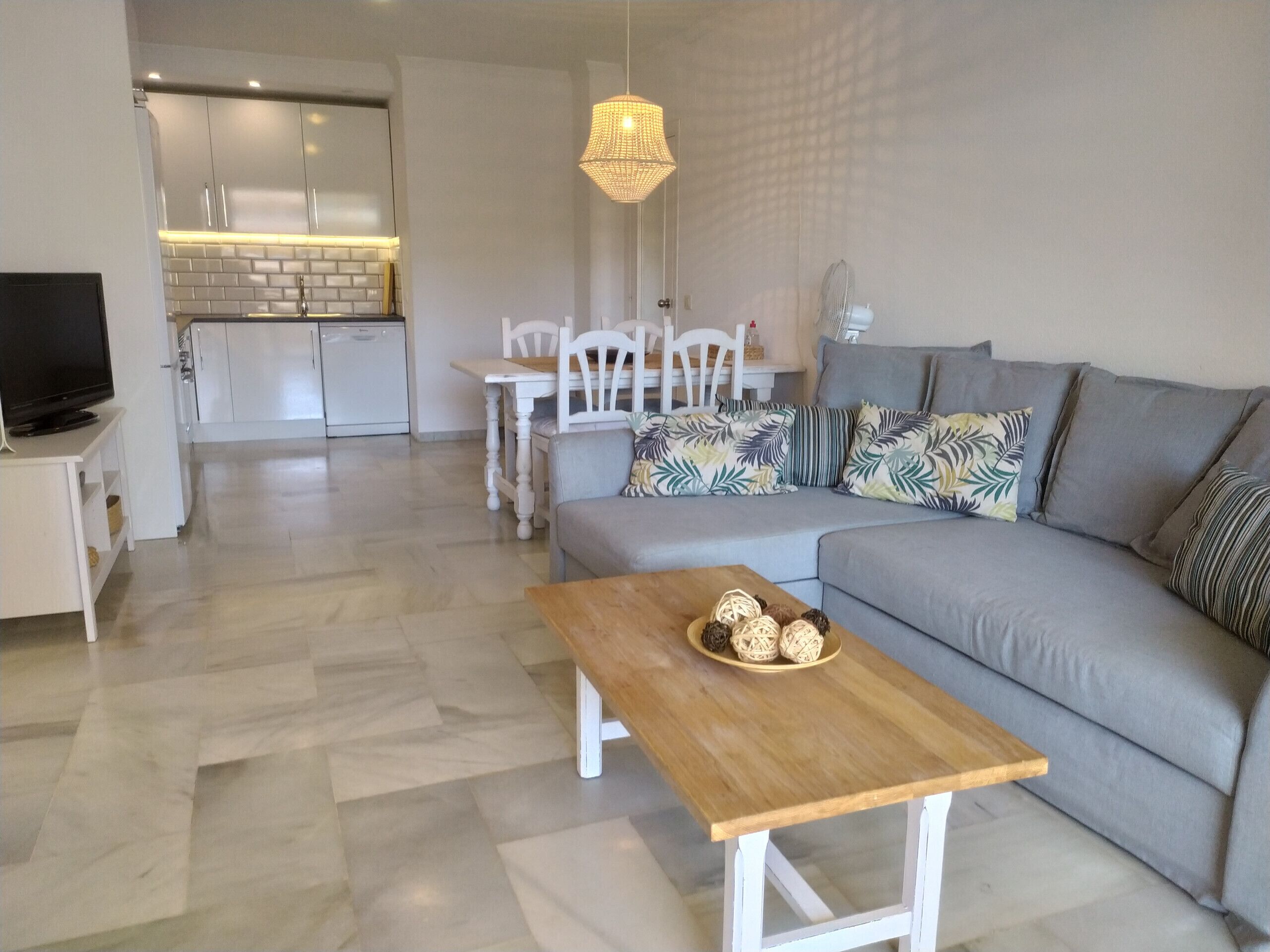A modern living room with a grey sofa, a wooden coffee table with decorative items, a TV on a stand, and a dining area with a white table and chairs.