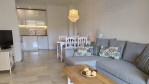 Ein modernes Wohnzimmer mit einem grauen Sofa, einem Holztisch mit Dekoration, einem Fernseher auf einem Schrank und einer Essecke mit weißem Tisch und Stühlen.