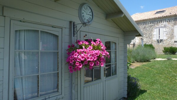 A house with a clock on the wall, a hanging flower basket, and a stone building in the background.