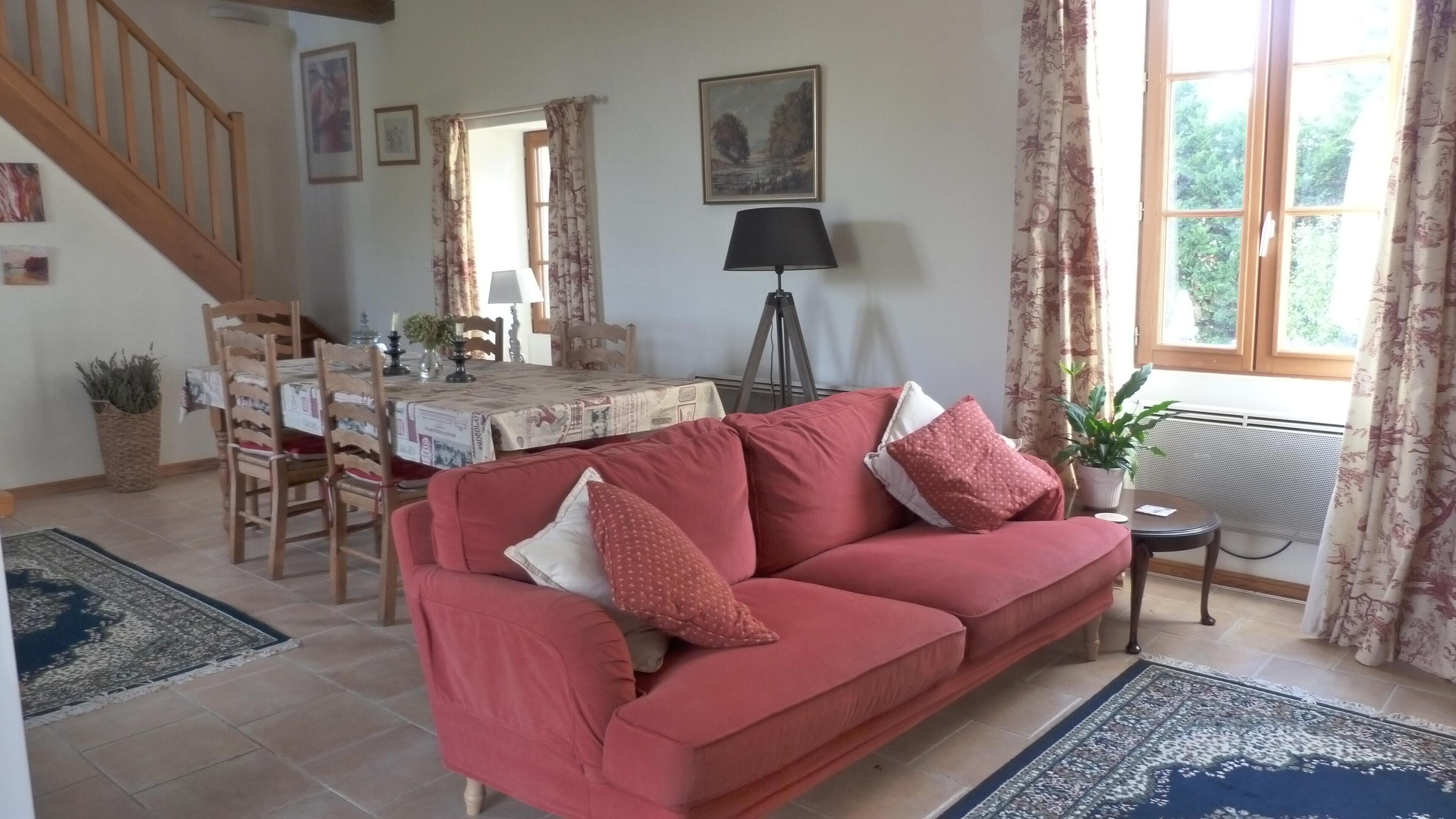 Een woonkamer met een rode bank, een eettafel, stoelen, een lamp en een raam met gordijnen.