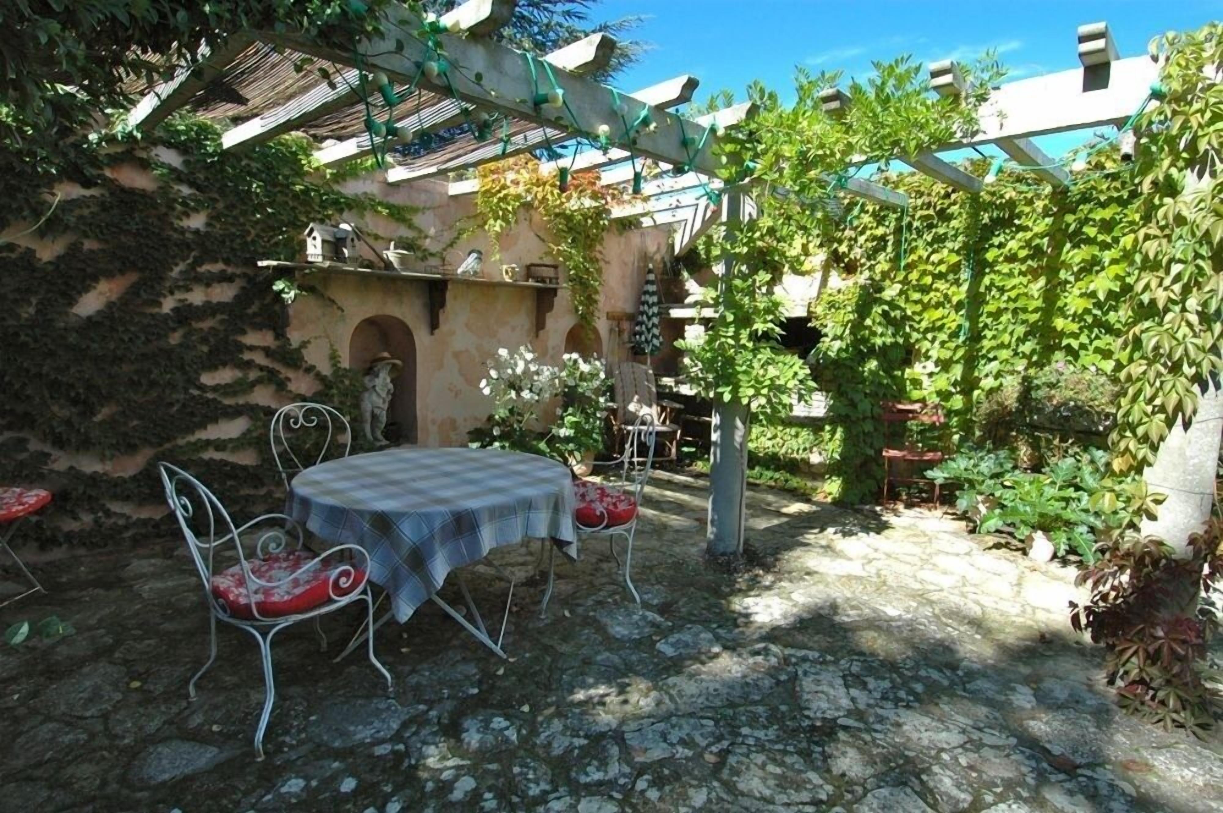 Une terrasse dallée de pierres, avec une table et des chaises, entourée de verdure et agrémentée d’une pergola.