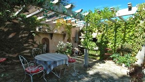 A stone-paved patio with a table and chairs, surrounded by greenery and a pergola.