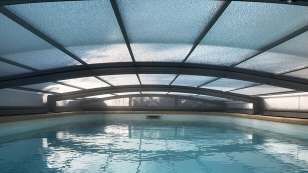 An indoor swimming pool with a glass ceiling, clear water, and a central diving board.