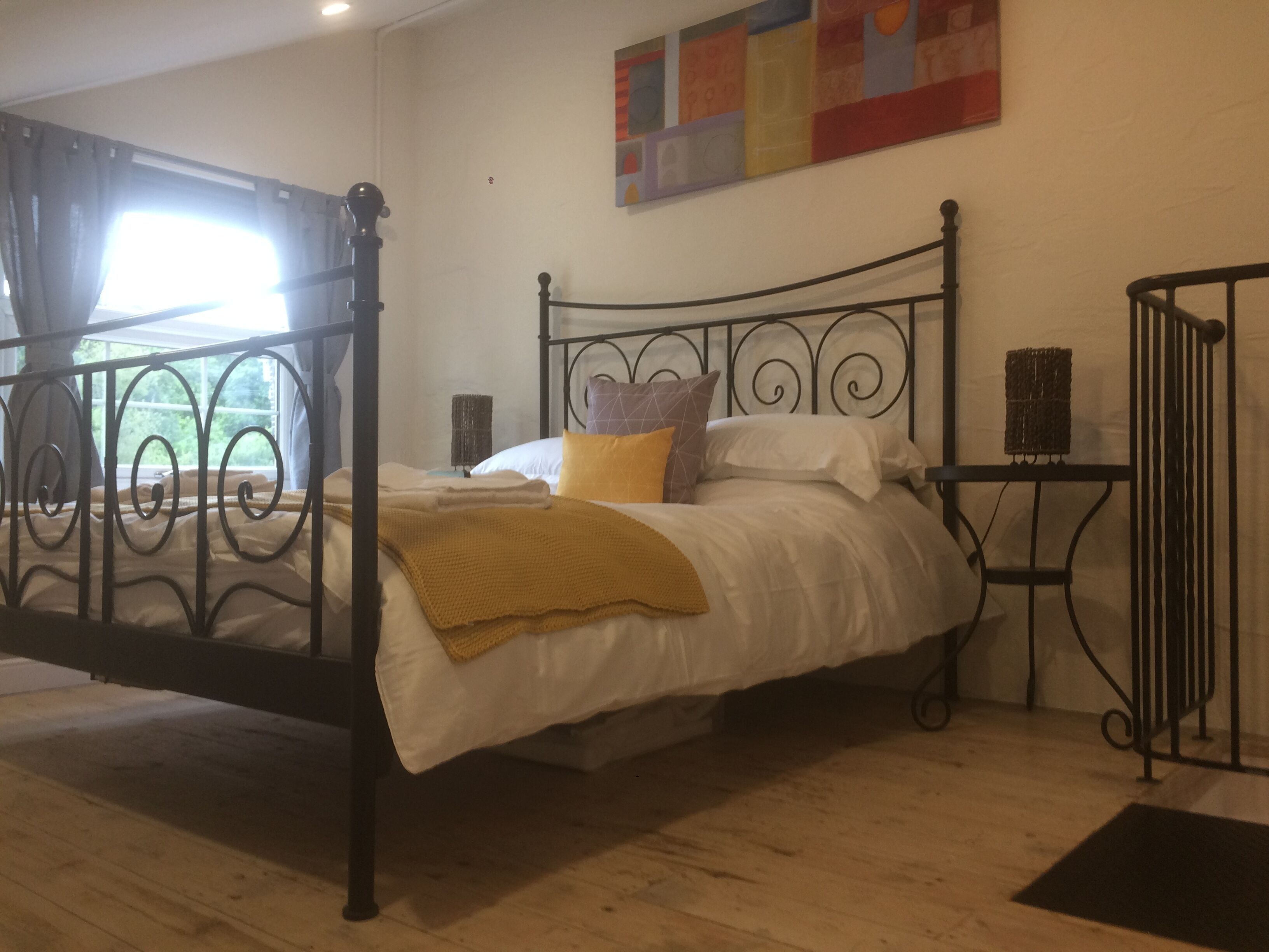 Quarto com estrutura de cama em metal, colcha branca e manta amarela. Há uma janela com cortinas, uma obra de arte na parede e uma pequena mesa com uma cesta.