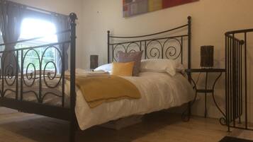 Quarto com estrutura de cama em metal, colcha branca e manta amarela. Há uma janela com cortinas, uma obra de arte na parede e uma pequena mesa com uma cesta.