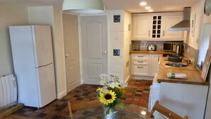 A compact kitchen with white cabinets, a refrigerator, and a dining table with a vase of flowers. - Cosy Cornish Bolt Hole, Pet Friendly (Truro)