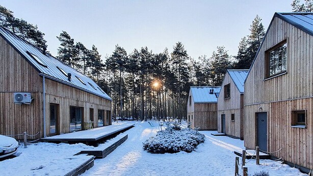 Ein mit Schnee bedecktes Holzgebäudeensemble mit einem zentralen Weg und umliegenden Bäumen.