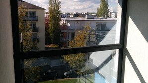 Vista attraverso una finestra su una zona residenziale con diversi edifici, tra cui una casa a due piani con balcone e un edificio moderno con tetto spiovente.