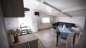 A compact kitchen with a gas stove, a sink, and a white refrigerator. There is a dining area with a table and chairs, and a dark-colored sofa. - Villa Barletta apartment mansarded in villa on the first floor number 5 (Marina di Acate)