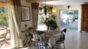 Un comedor con mesa y sillas, una araña, un florero con flores y vista a la cocina a través de una puerta abierta.