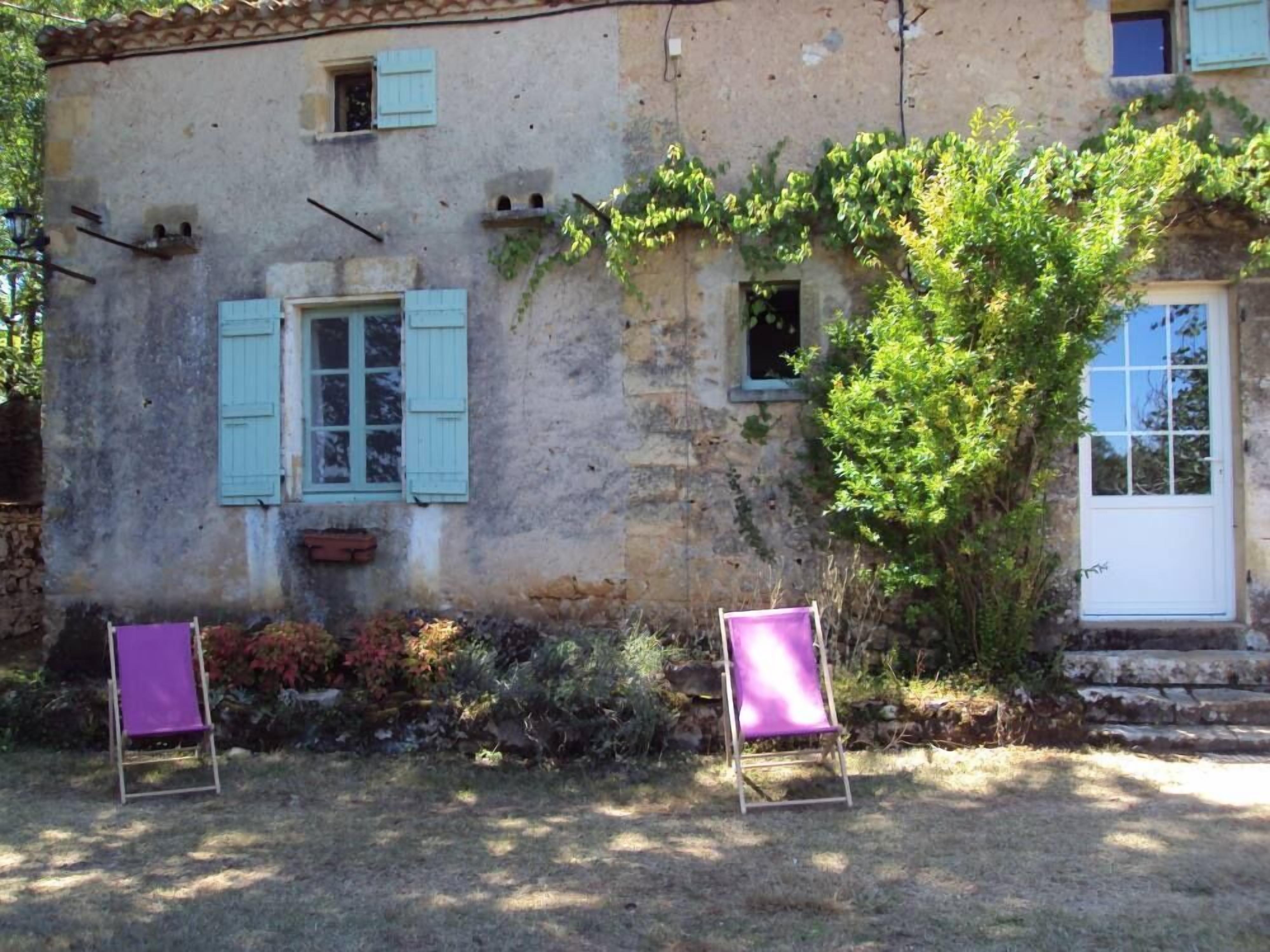 A rustic stone building with two windows and a door, surrounded by greenery and a small garden area.