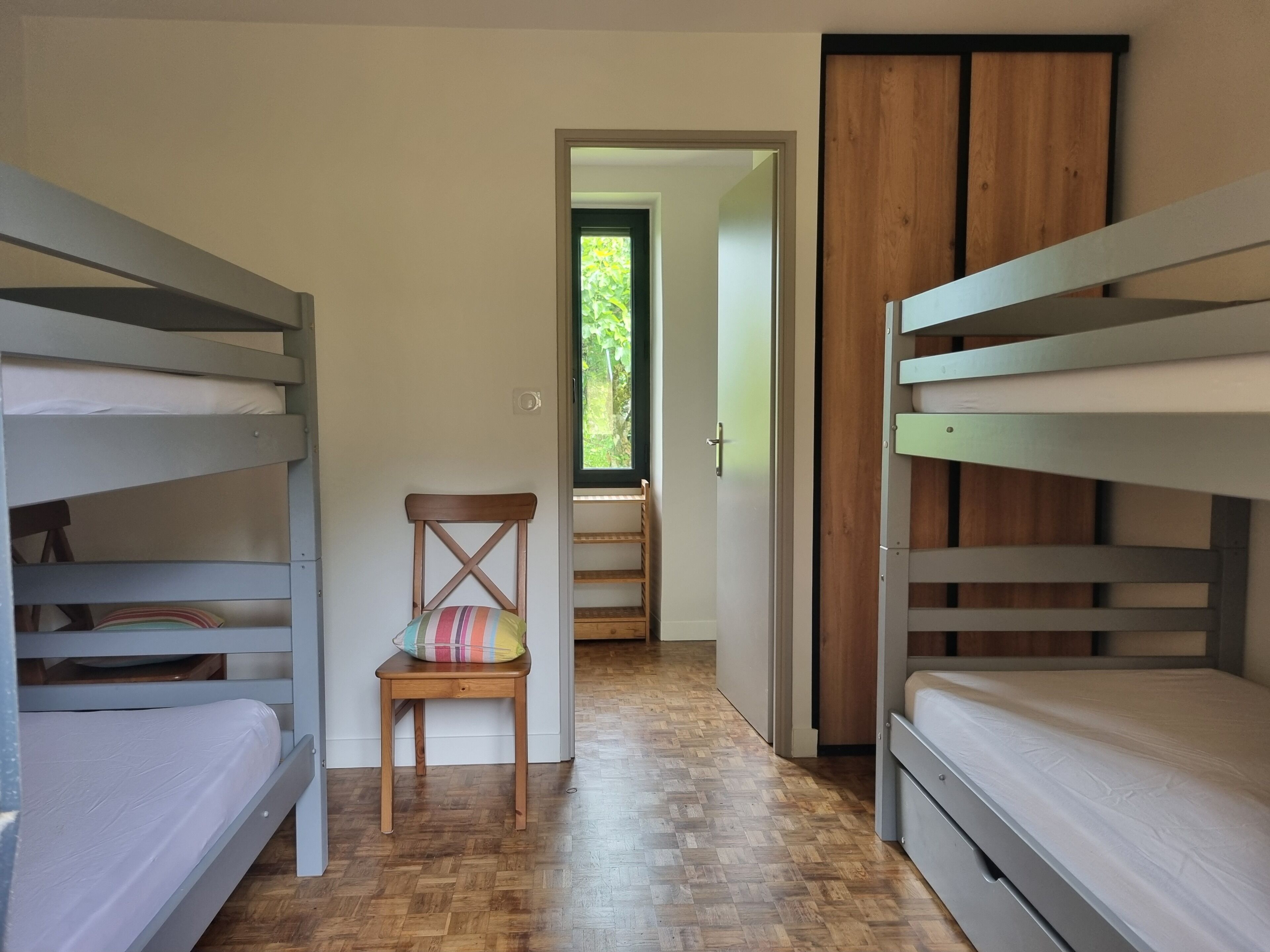 A room with bunk beds, a wooden chair, a window with a view of greenery, and a door leading to another room.