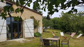 A rustic house with a large sliding glass door, a small patio with a table and chairs, and a grassy yard with a few potted plants.
