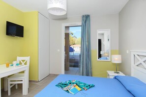 A hotel room with a bed, a desk, a chair, a TV, a window with curtains, and a view of an outdoor swimming pool. - Apartment in France with Pool and Pine Views (La Londe les Maures)