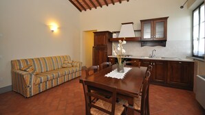 A compact living space with a dining table, wooden chairs, a striped sofa, and a kitchen area with wooden cabinets and a window.