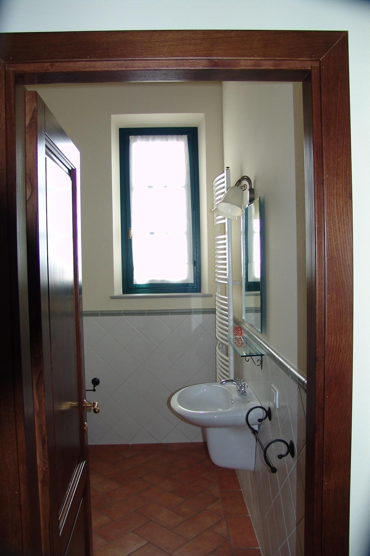 A bathroom with a wooden door, a window with a white blind, a white sink with a mirror above it, and a towel rack.