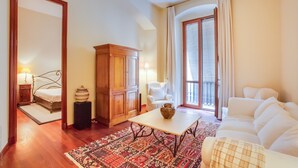 Una habitación de hotel con una cama, un armario de madera, un sillón blanco, un sofá, una mesa con un jarrón y una alfombra estampada.