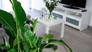 A modern living room with a white TV stand, a flat-screen TV, a white coffee table, and a large green plant in the foreground. - Modern apartment in Puerto marina (Benalmádena)