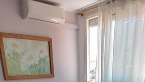 A bedroom with a bed, a nightstand, a window with curtains, an air conditioning unit, and a painting on the wall.