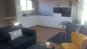 Un espacio habitable compacto con sofá, un sillón amarillo, una mesa de centro de vidrio, una mesa de comedor con sillas y una kitchenette con estufa y horno.