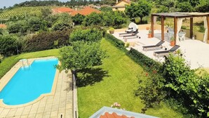 Ein Swimmingpool mit Fliesenveranda, eine Pergola mit Sitzgelegenheiten und ein Blick auf Weinberge im Hintergrund.