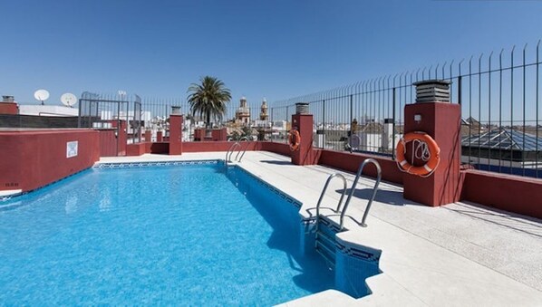 A rooftop swimming pool with a clear view of the city skyline, a palm tree, and a lifebuoy on the railing.