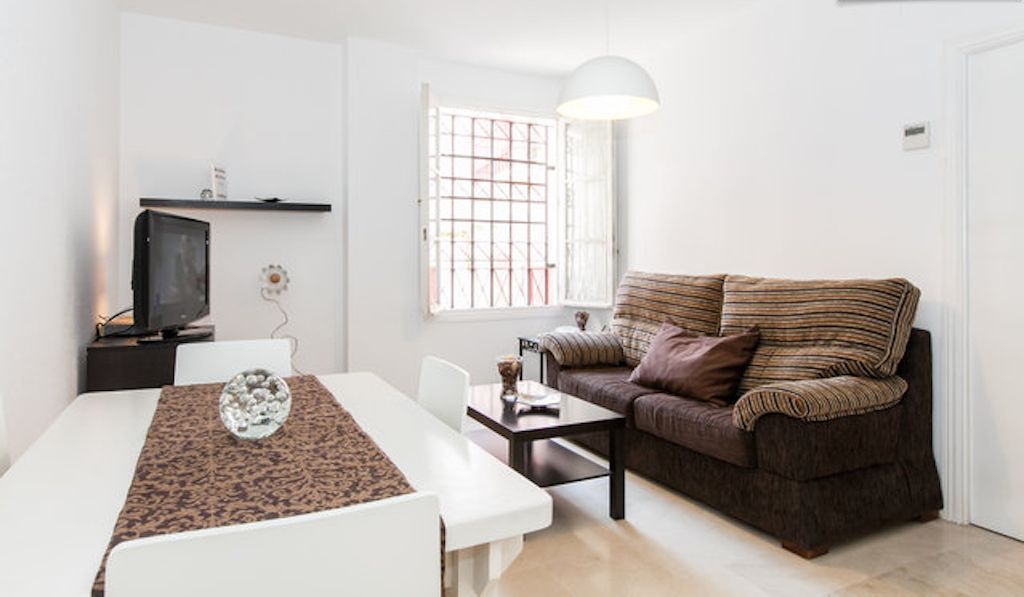 A modern living room with a white sofa, a dining table with a patterned table runner, a television on a stand, and a large window with a grid pattern.