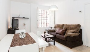 Una sala de estar moderna con un sofá blanco, una mesa de comedor con mantel decorado, un televisor sobre un mueble bajo y un ventanal grande con diseño de cuadrícula.