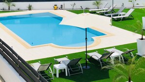 Una piscina con un deck circundante, sillas de descanso y una pequeña zona ajardinada.