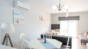 Un comedor con una mesa blanca, sillas y un centro de mesa con velas azules. Se observa un aire acondicionado blanco en la pared, un cuadro y un televisor al fondo.