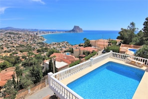 Uma piscina com balaustrada branca, rodeada por uma área residencial com casas de telhado vermelho e vista para o mar e uma formação rochosa imponente.