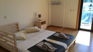 Ein Schlafzimmer mit einem hölzernen Bett, einem Nachttisch mit Lampe, einem Kleiderständer und Blick ins Freie durch ein großes Fenster.