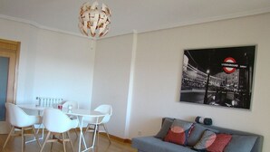 Ein modernes Wohnzimmer mit einem grauen Sofa, weißen Stühlen und einem Esstisch. Eine besondere, runde Leuchte hängt von der Decke, und ein U-Bahn-Poster ziert die Wand.