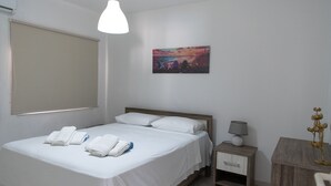 Un lit bien fait, avec du linge de lit et des serviettes blanches, une table de chevet avec une lampe et un tableau représentant un coucher de soleil accroché au mur.