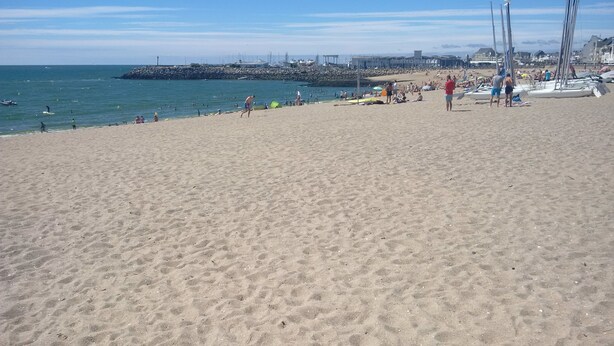 A sandy beach with people, boats, and a pier in the background.