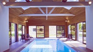 A modern indoor swimming pool with clear glass walls, wooden ceiling beams, and a stone wall in the background.