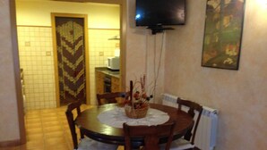 Un comedor con una mesa redonda, cuatro sillas, un centro de mesa decorativo, un televisor montado en la pared y piso de baldosas.