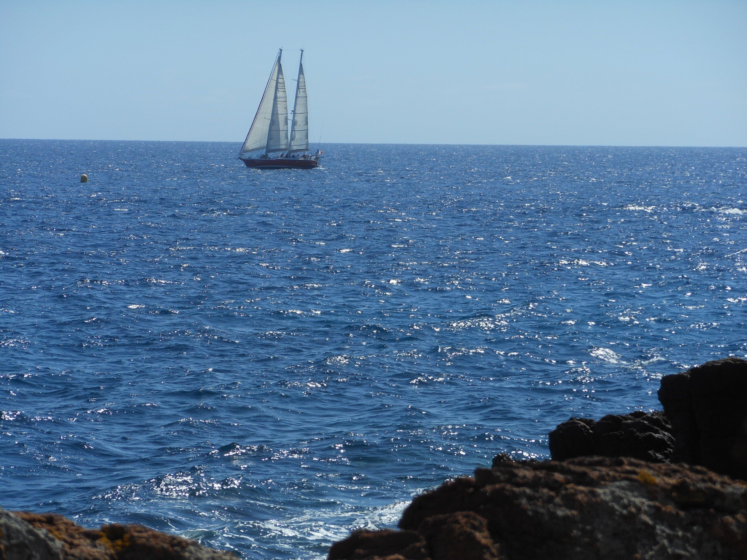 A sailboat on a calm sea with rocky coastline in the foreground.