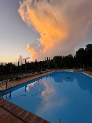 Une piscine avec des chaises longues, donnant sur des arbres et un ciel au coucher du soleil.