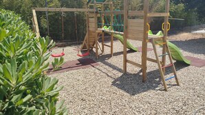 A wooden playground structure with swings, slides, and climbing elements, surrounded by gravel and greenery.