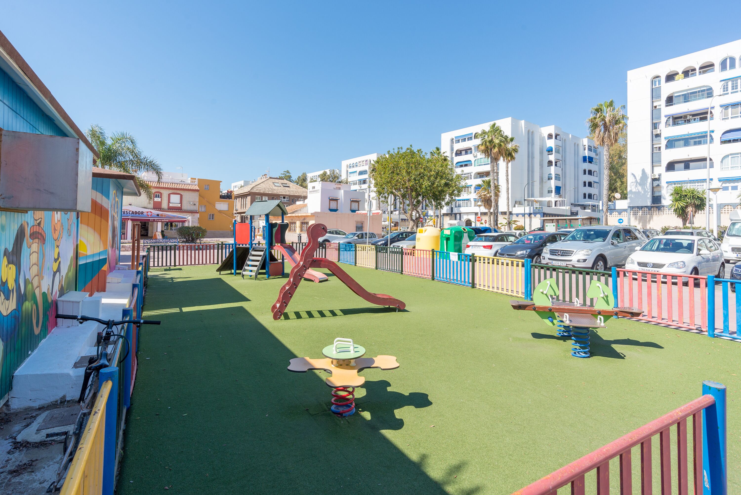 A playground with a slide and swings, surrounded by a fence, with buildings and parked cars in the background.