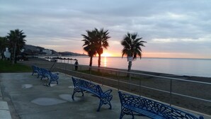 Ein Strand mit blauen Metallbänken, Palmen und einem Sonnenuntergang über dem Wasser.