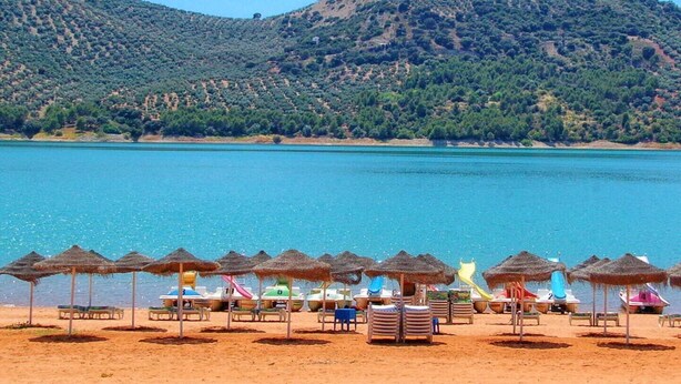 A beach with thatched umbrellas and lounge chairs, a clear blue sea, and a hilly landscape in the background. - Apartment with big terrace and fantastic views (Iznajar)