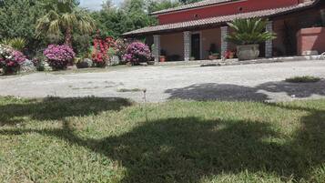 A house with a red facade, stone pillars, and a tiled roof. There are flowering plants and a palm tree in the garden.
